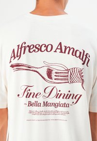 Cremefarvet bomulds t-shirt med maroon tekst og gaffelgrafik, med ordene "Alfresco Amalfi," "Fine Dining," og "Bella Mangiata."