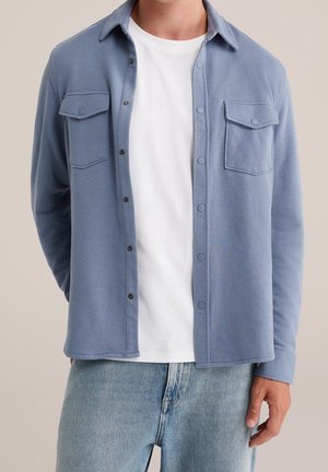 Mand iført lyseblå skjorte med knapper og brystlommer over hvid T-shirt og lyseblå denimjeans, stående mod en neutral baggrund.