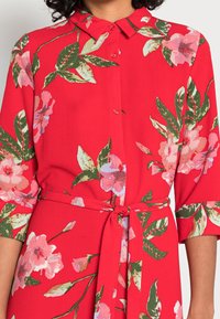 Robe fleurie rouge à manches trois-quarts, boutonnée à l'avant, avec ceinture à nouer à la taille. Motif de fleurs roses et feuilles vertes.