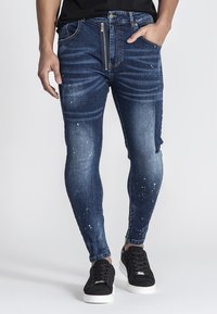 Jeans de denim ajustados en azul oscuro con un lavado desgastado, ligeras áreas destruidas y un panel contrastante en una pierna. Cuenta con cierre de cremallera.