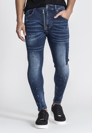 Slim-fit denim jeans in dark blue with a faded wash, light distressed spots, and a contrasting panel on one leg. Features a zip-fly closure.