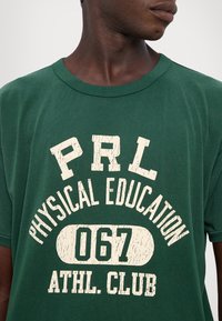 Zelené bavlněné tričko s kulatým výstřihem. Obsahuje bílej opotřebovaný text: "PRL PHYSICAL EDUCATION 067 ATHL. CLUB." Uvolněný střih.