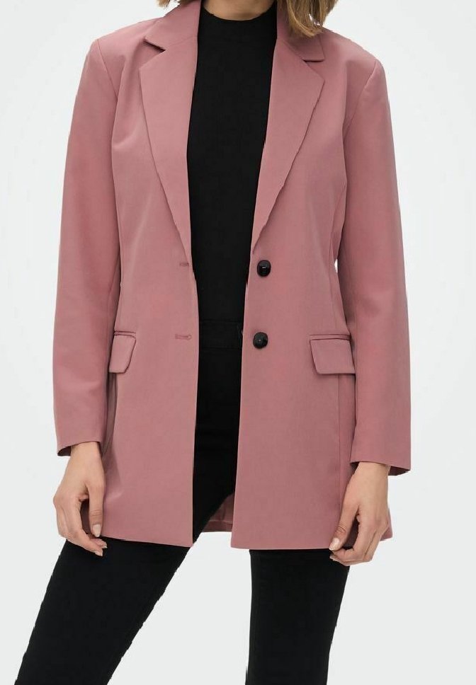 Femme portant un blazer rose poussiéreux avec des boutons noirs sur un haut noir et un pantalon noir, debout devant un fond clair uni.