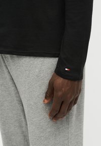 Persona dalla pelle scura che indossa una camicia nera a maniche lunghe e pantaloni grigi, mano rilassata lungo il fianco, piccolo logo rosso, bianco e blu sul polsino della manica.
