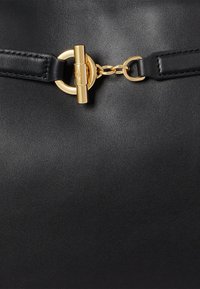 Fekete bőrtáska arany kör alakú fémrészlet dísszel és láncszem részlettel, sima textúrával és minimalista dizájnal.