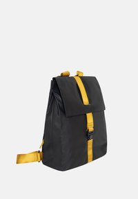 Zaino nero in nylon con accento giallo sulla tracolla, forma rettangolare, chiusura con patta anteriore e spallacci regolabili.