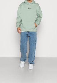 Felpa verde menta con tasca frontale e logo corsivo nero. Indossata con jeans blu chiaro a gamba dritta e sneakers bianche.