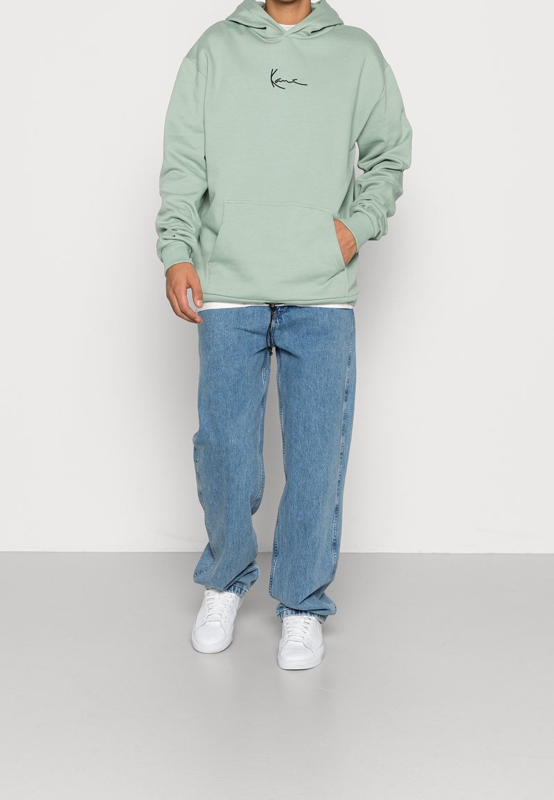 Felpa verde menta con tasca frontale e logo corsivo nero. Indossata con jeans blu chiaro a gamba dritta e sneakers bianche.