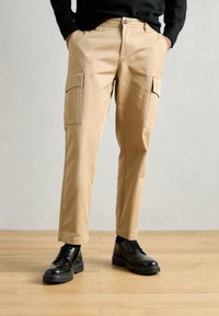 Beige cargo pants with two large side pockets, button closure, and slim fit; paired with black lace-up shoes on a wooden floor.