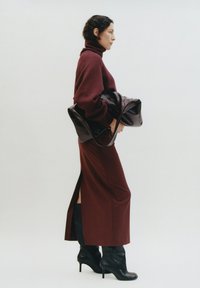 Bordeaux coltrui, een aansluitende lange rok met een zijnaad, en zwarte hakken. Model houdt een donkere leren tas vast. Minimalistische achtergrond.