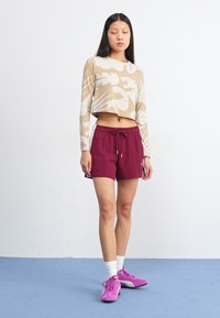 Une jeune femme se tient sur un sol bleu, portant un crop top avec un design beige et blanc, un short bordeaux et des baskets roses.