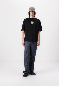 Black t-shirt with a small, colorful design at the chest; loose grey cargo pants with multiple pockets; black shoes; grey cap.