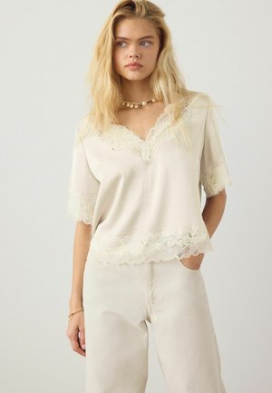 Blond vrouw draagt een crèmekleurige blouse met V-hals afgewerkt met kant en bijpassende broek met hoge taille, met een halsketting en armband met gouden munten.