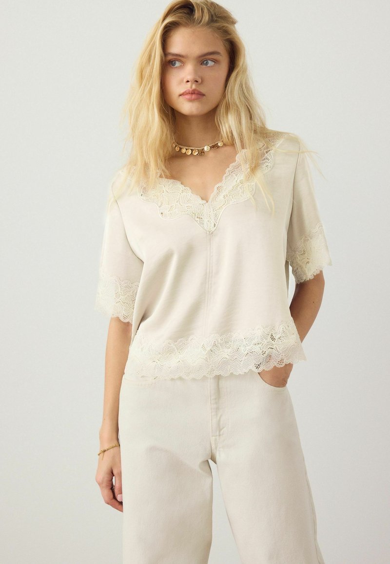Femme blonde portant un chemisier crème à col en V orné de dentelle et un pantalon taille haute assorti, avec un collier et un bracelet en pièces d'or.