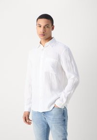 Weißes Leinenhemd mit langen Ärmeln, Knopfkragen, einer einzelnen Brusttasche und strukturiertem Stoff. Getragen mit hellblauen, zerrissenen Jeans.