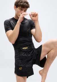 Μαύρο σετ αθλητικής ένδυσης που περιλαμβάνει ένα υφασμάτινο κοντομάνικο τοπ και σορτς με μοτίβο. Η μέση έχει το λογότυπο "VENUM" σε χρυσό, με λογότυπο λιονταριού.