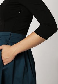 Top negro ajustado con mangas tres cuartos, combinado con una falda verde azulado con un diseño plisado y bolsillo lateral.