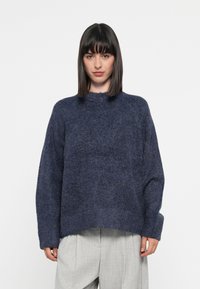Mørkeblå, oversized genser laget av teksturert stoff med høy hals og vide ermer. Sømmen er litt cropped.