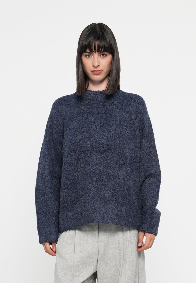 Mørkeblå, oversized genser laget av teksturert stoff med høy hals og vide ermer. Sømmen er litt cropped.