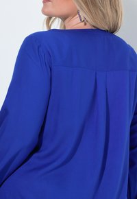 Blusa blu con una texture liscia, caratterizzata da un design con schiena plissettata e un collo semplice. Maniche lunghe con vestibilità comoda.