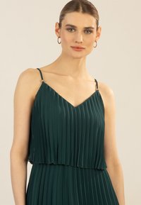 Plissiertes dunkelgrünes Kleid mit V-Ausschnitt, verstellbaren Spaghettiträgern und silbernen Hardware-Details an den Trägern.