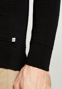 Detail černého pleteného svetru s žebrovanou texturou a přiléhavými manžetami, s malou bílou štítkem poblíž bočního švu.