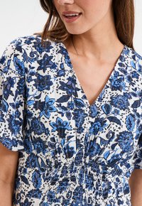 Bluse mit blauem Blumenmuster, V-Ausschnitt, elastischem Bund und kurzen Ärmeln. Der Stoff hat eine weiche Textur und ein detailliertes Muster.