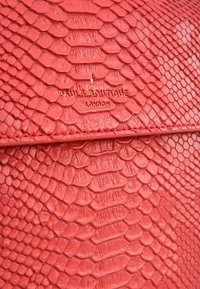 Červená kabelka z imitace hadí kůže s texturovaným povrchem a zlatým embosovaným logem. Design má štíhlý, protáhlý tvar s klopovým uzávěrem.