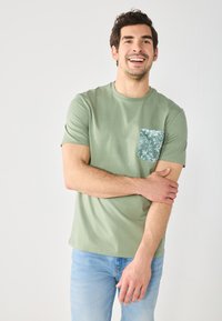 Green cotton t-shirt with a round neckline and short sleeves. Features a patterned blue pocket. Worn with light blue jeans.