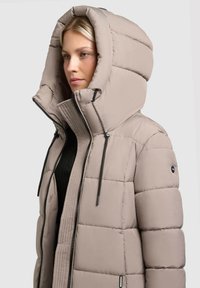 Puffjacket i lys beige med en stor hætte, vertikal quilting, lynlås foran og sorte snore detaljer. Tekstureret indre foring.