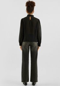 Zwarte, gestructureerde blouse met pofmouwen en een gerimpelde kraag; detail met sleutelgat aan de achterkant. Gecombineerd met losse, donkergrijze jeans.