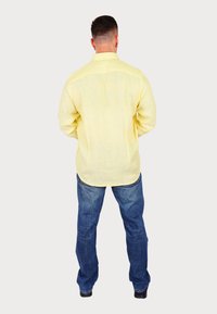 Camicia giallo chiaro a maniche lunghe in lino, abbinata a jeans blu in denim. La camicia ha uno spacco posteriore ed è lasciata fuori dai pantaloni.