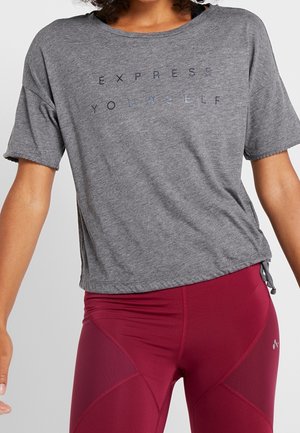 Nainen pukeutuneena harmaaseen lyhythihaiseen paitaan, jossa lukee "EXPRESS YOURSELF", ja tiukkoihin, tummanpunaisiin urheilutrikooihin.