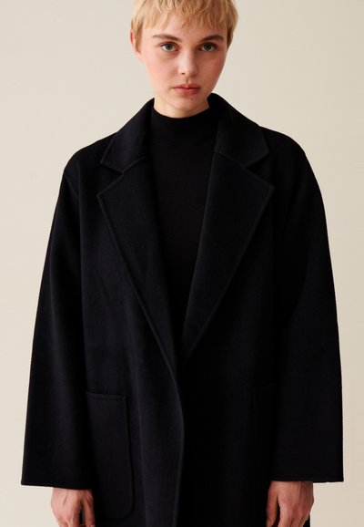 Cappotto di lana nero con colletto a punta, due tasche frontali e vestibilità ampia. Il tessuto ha una texture liscia. Il modello indossa un dolcevita nero sotto.