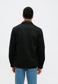 Fekete dzseki strukturált gallérral, gombos mandzsettával és sima textúrával. Halványkék farmernadrág fölött viselve.