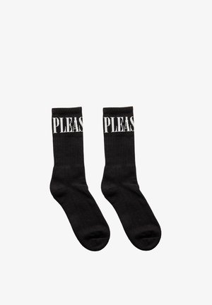 Para czarnych skarpet sportowych z białym napisem "PLEASE" blisko górnego ściągacza, ułożonych płasko na białym tle.
