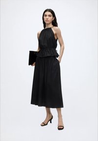 Γυναίκα με μαύρο κορσέ χωρίς μανίκια και μαύρο φαρδύ παντελόνι μέχρι το μέσο της γάμπας, κρατώντας μαύρο τσαντάκι και φορόντας μαύρες γόβες.