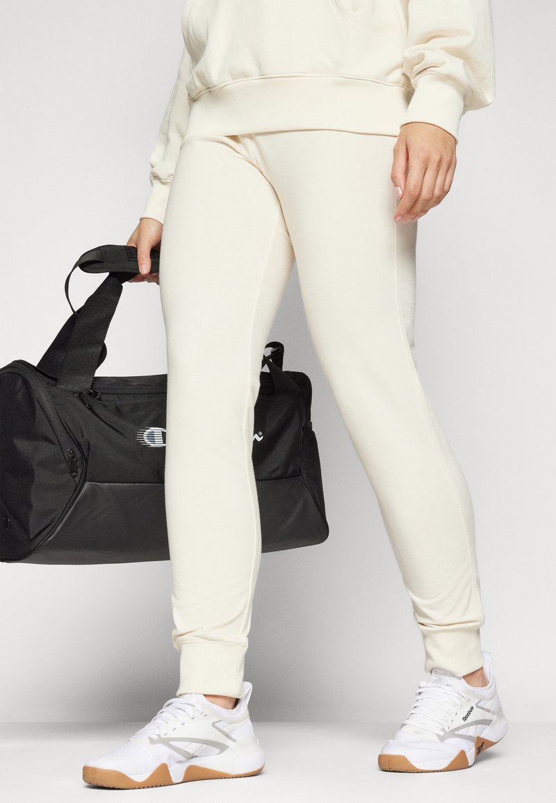 Joggers color crema con un diseño ajustado, puños acanalados y una textura suave, combinados con una bolsa de deporte negra y zapatillas blancas.