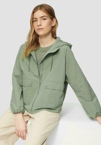 Veste à capuche vert clair en matériau léger, dotée d'une fermeture éclair à l'avant, de deux rabats de poche et de poignets élastiques, portée sur un pull gris.