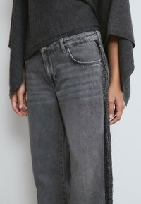 Persona vistiendo jeans de mezclilla grises con detalles de flecos negros en las costuras laterales y un suéter de punto gris oscuro con mangas anchas.