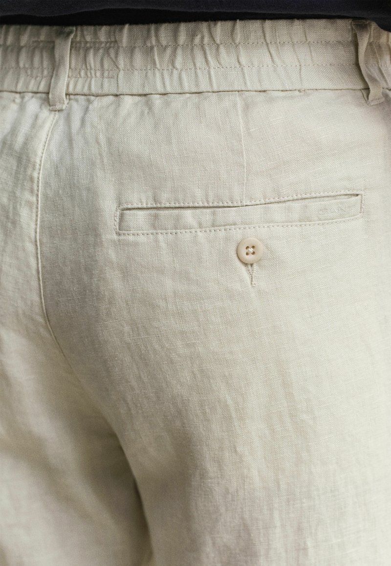 Close-up van beige linnen broek met elastische tailleband, achterzak met paspel en knoop en zichtbare riemlussen.