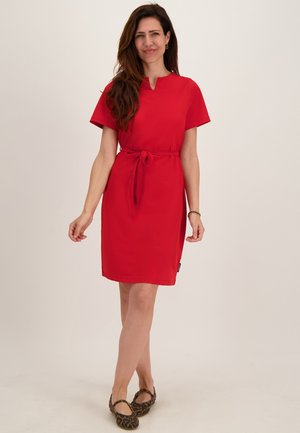 Vrouw staat en lacht, draagt een knielange rode jurk met korte mouwen, een taillekoord en platte schoenen met luipaardprint.