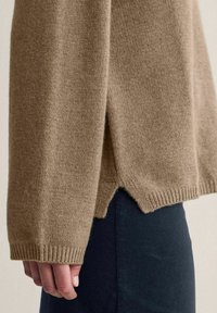 Beige neulepaita, jossa on ribbireunat ja korkea-alhainen helma, jossa on sivurei'ät. Pehmeä rakenne ja rento istuvuus.