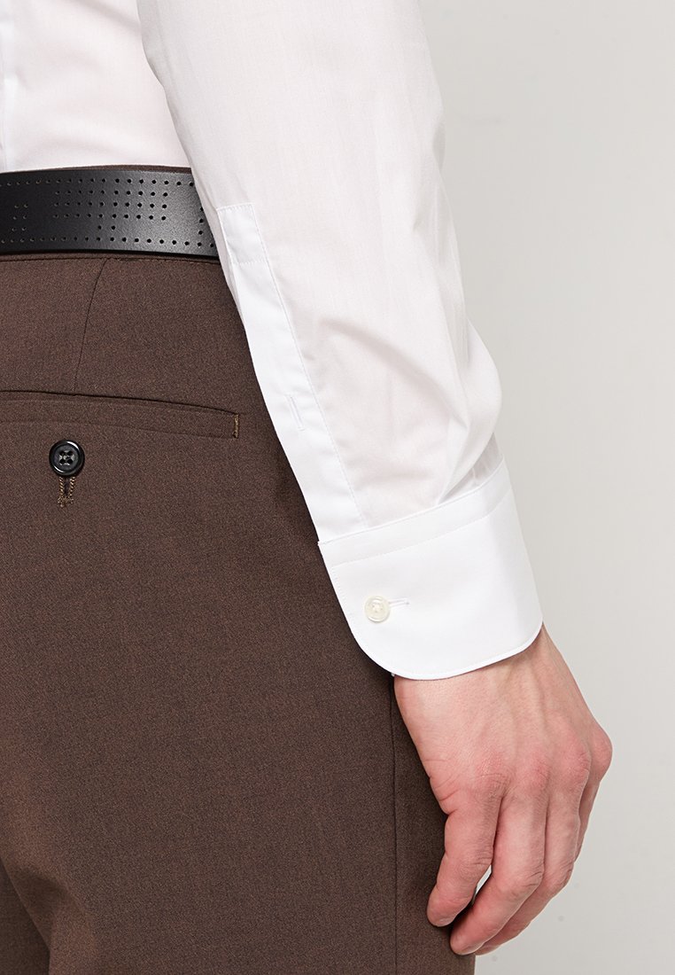 Camisa blanca de mangas largas con puños abotonados, combinada con pantalones marrones que tienen un bolsillo trasero y un cinturón negro perforado.