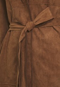 Vêtement en daim marron avec une ceinture nouée, légèrement texturé, coutures apparentes. Matériau doux et lisse avec une finition mate.