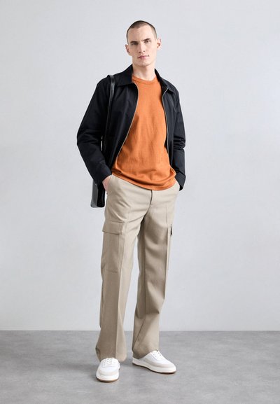 Veste zippée noire sur un t-shirt orange, associée à un pantalon cargo beige. Baskets blanches avec une semelle texturée. Design simple et moderne.