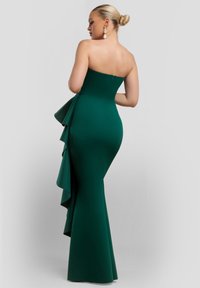 Vihreä, olkaimeton mekko, jossa on istuva yläosa ja liehuva helma. Mekossa on sivulla kerroksittainen röyhelökoriste. Materiaali näyttää sileältä ja joustavalta.