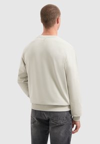Mann, der einen hellbeigen Pullover und dunkle graue Jeans trägt, von hinten vor einem schlichten hellen Hintergrund betrachtet.