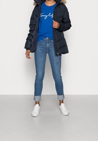 Granatowa kurtka puchowa z futrzanym kapturem, niebieska koszulka z białym napisem, jasnoniebieskie rurki i białe sneakersy.