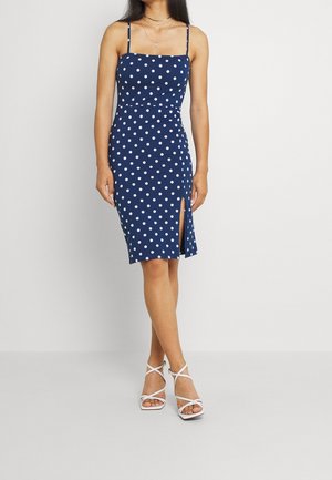 Femme portant une robe sans manches bleu marine à pois blancs avec une fente à l'avant, associée à des sandales blanches à talons hauts et lanières.